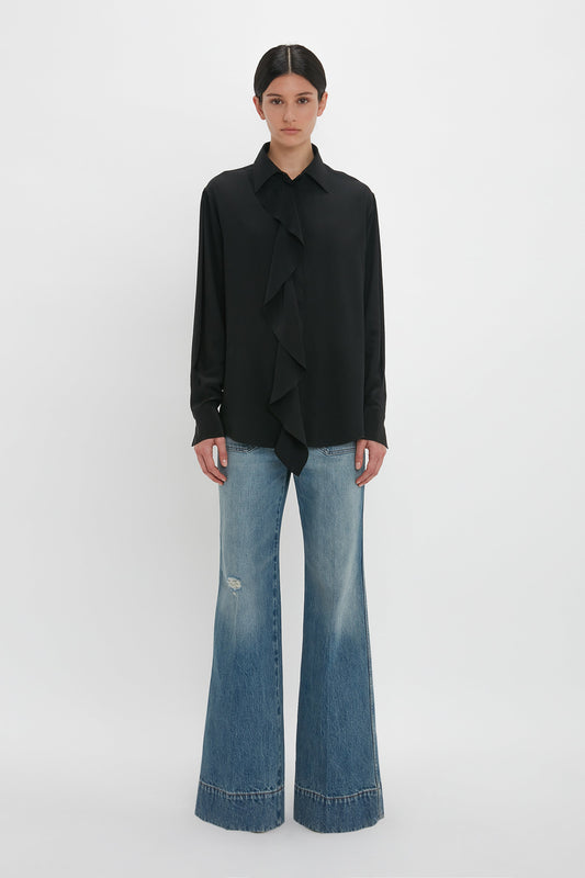 A woman stands against a white background, wearing a Victoria Beckham black silk blouse with an asymmetric ruffle and wide-legged denim jeans.