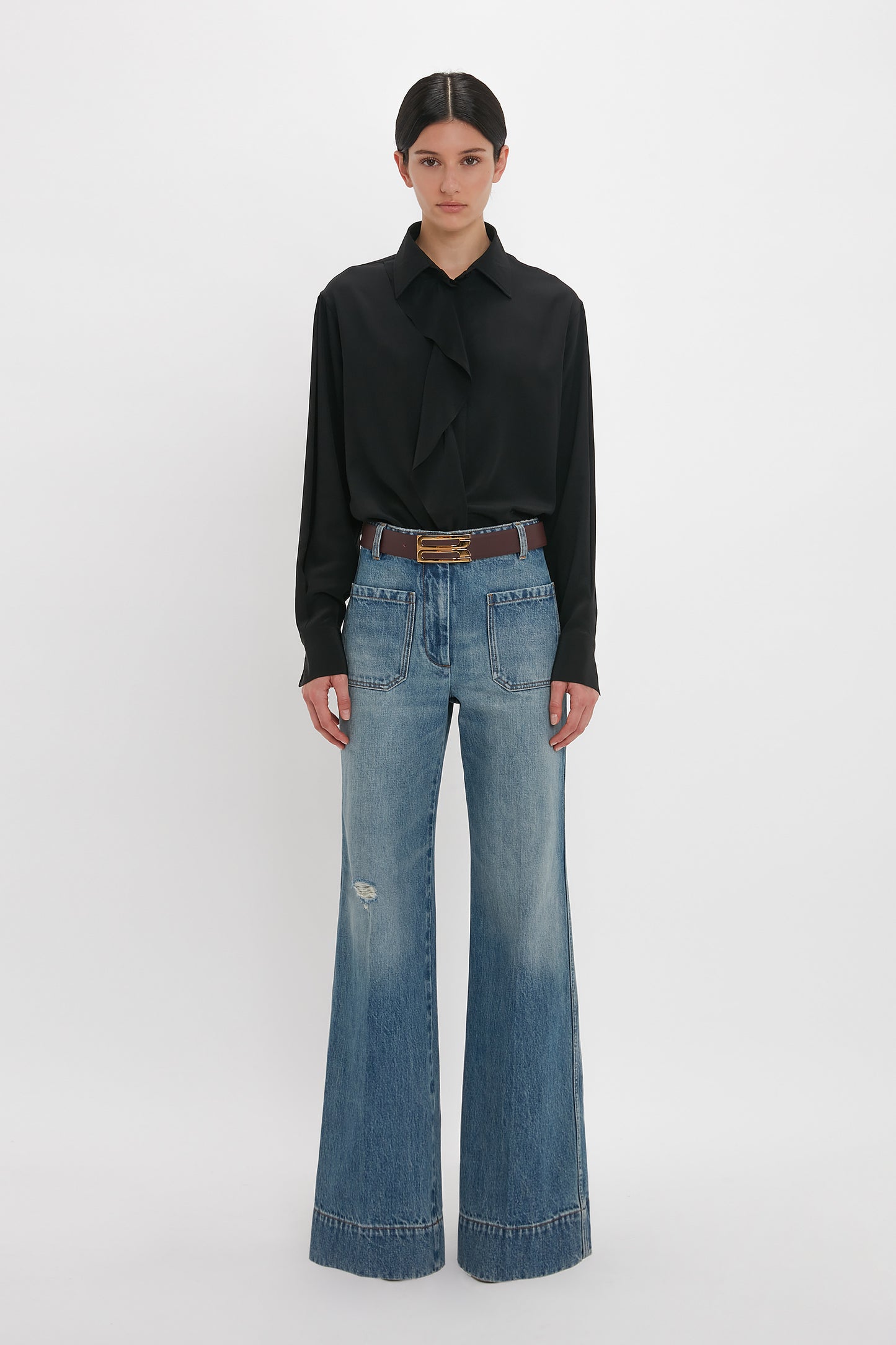 Person wearing a black long-sleeve blouse with a bow tie and wide-leg Victoria Beckham Alina High Waisted Jean In Worn Blue Wash featuring seventies-inspired patch pockets, standing against a plain white background.