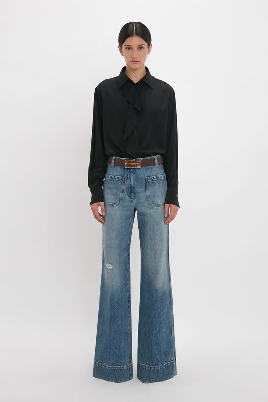 A woman stands against a white background wearing a Victoria Beckham black asymmetric ruffle blouse and distressed wide-leg jeans, accessorized with a brown belt.