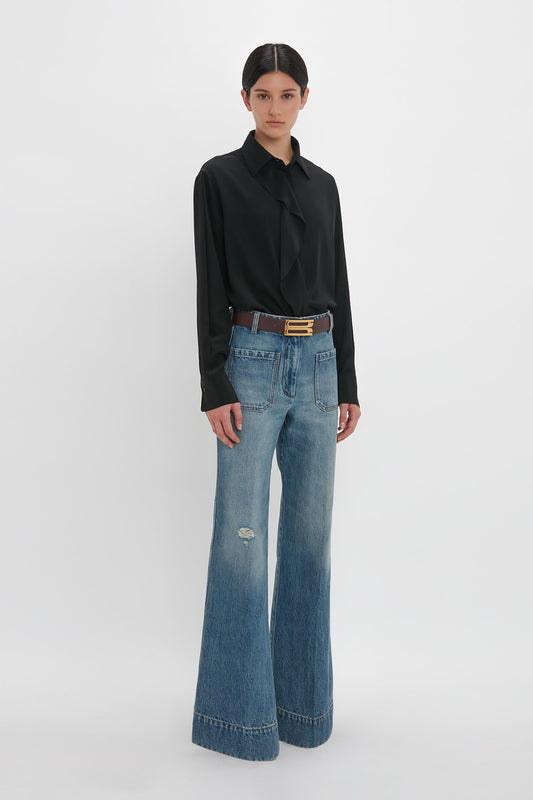 A woman stands against a white background, wearing a Victoria Beckham black silk blouse and distressed blue jeans with a brown belt.