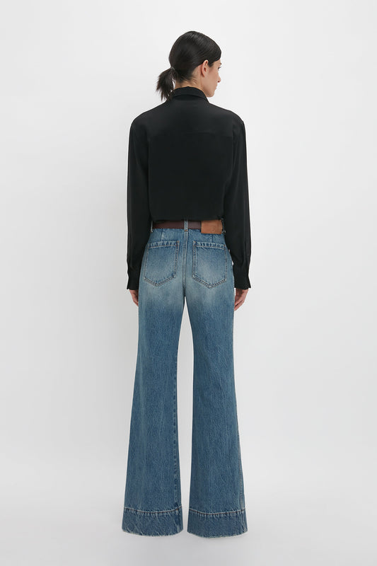 A woman viewed from behind wearing a Victoria Beckham asymmetric ruffle blouse in black and blue flared jeans with a brown belt on a white background.