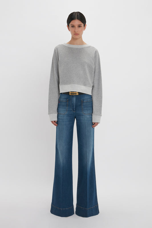 Woman in Victoria Beckham grey marl cropped sweatshirt and wide-leg blue jeans standing against a white background.