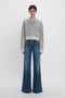 Woman in Victoria Beckham grey marl cropped sweatshirt and wide-leg blue jeans standing against a white background.