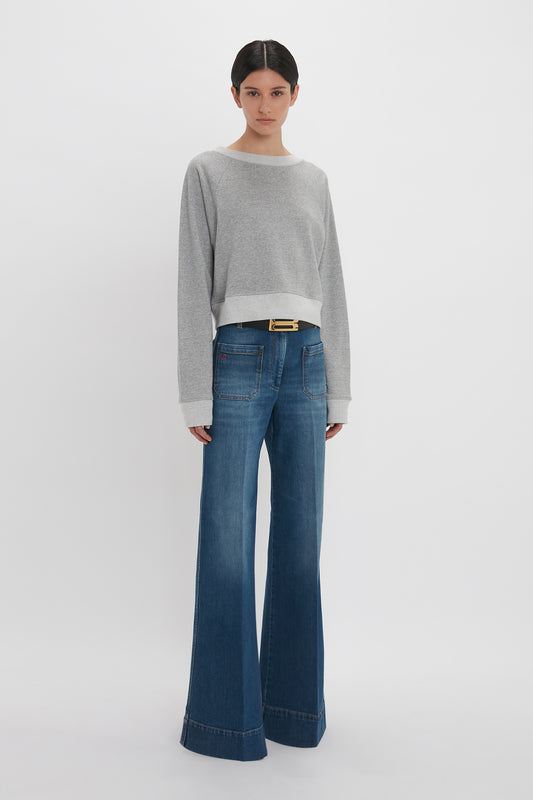 A woman wearing a Victoria Beckham Cropped Sweatshirt in Grey Marl and blue flared jeans standing against a white background.
