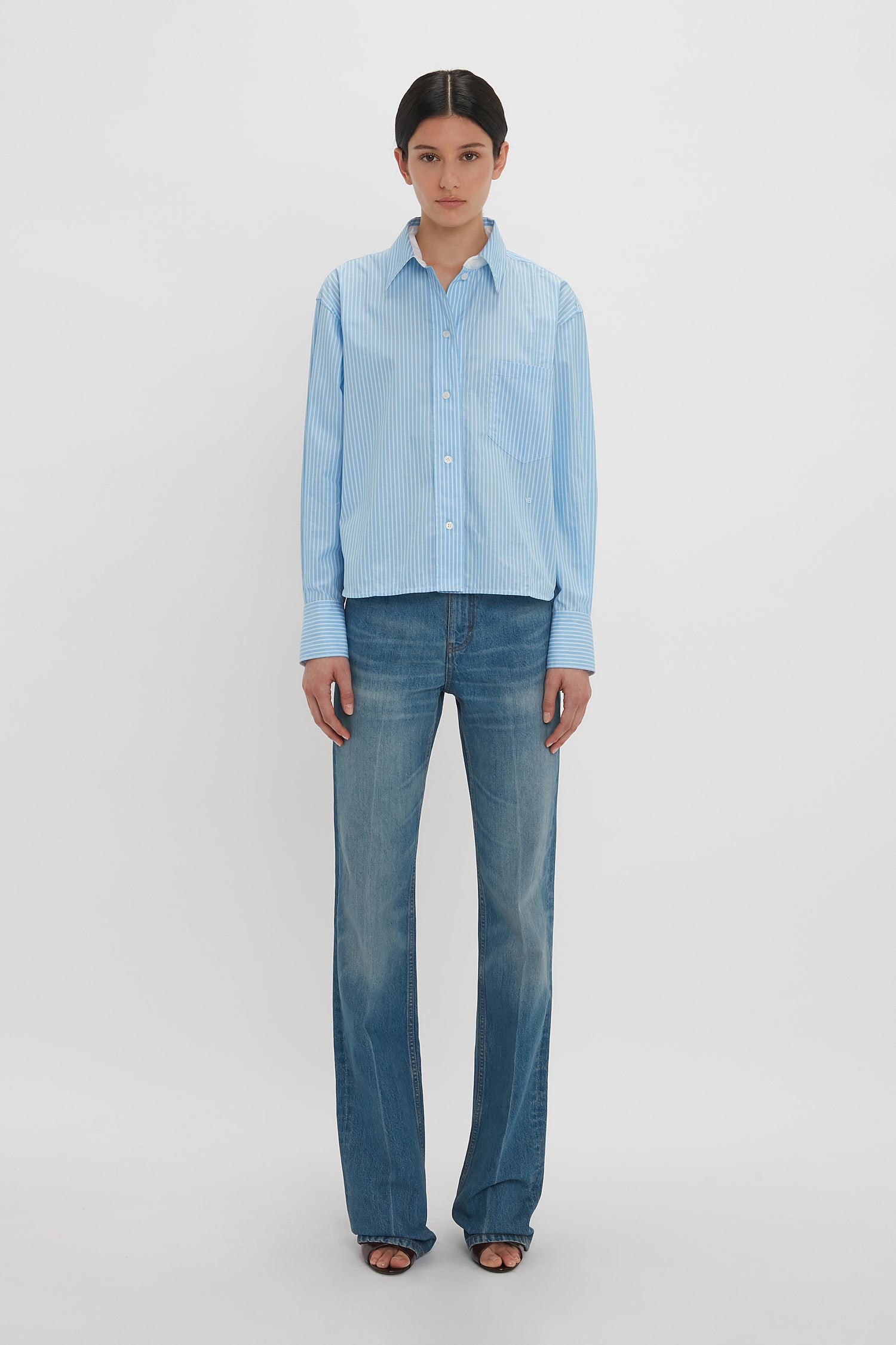 Person standing against a plain background, wearing a Cropped Long Sleeve Shirt In Marina-White by Victoria Beckham, blue jeans, and black open-toe shoes.