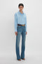 Person in a Cropped Long Sleeve Shirt In Marina-White by Victoria Beckham, blue jeans, brown belt, and black sandals standing against a plain white background.