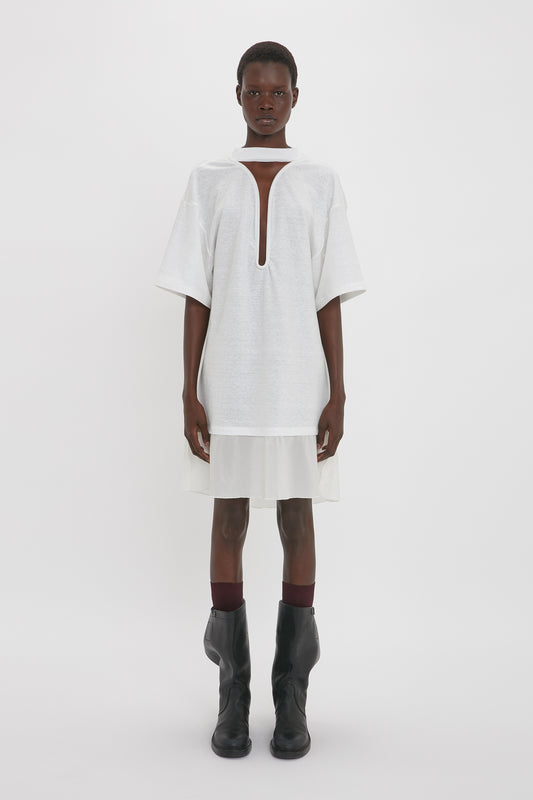 A person stands against a plain white background, wearing the Victoria Beckham Frame Cut-Out T-Shirt Dress In White, complemented by a white skirt underneath and black boots.