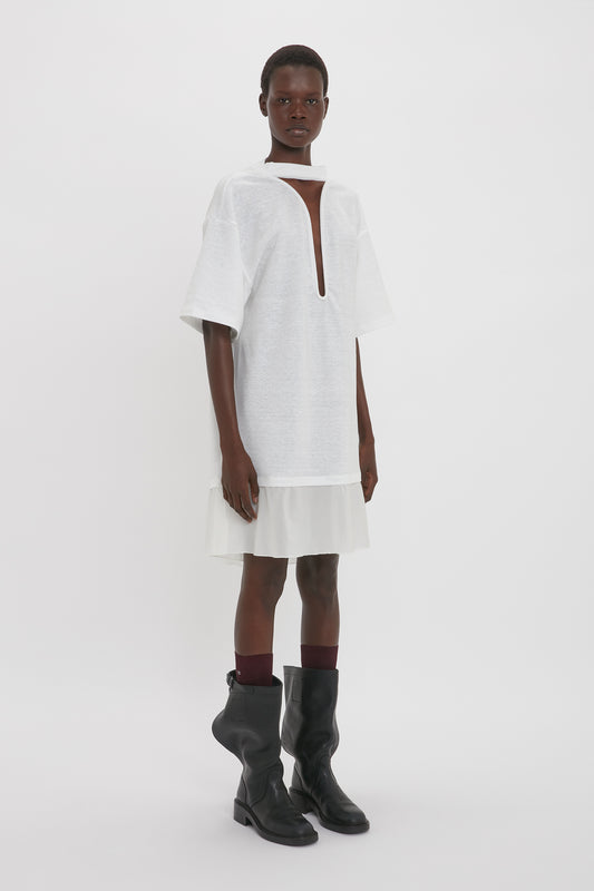 A person wearing a white Victoria Beckham Frame Cut-Out T-Shirt Dress In White with a keyhole neckline and black boots stands against a plain white background.