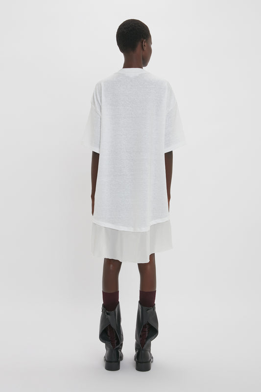 A person stands facing away, wearing an oversized Victoria Beckham Frame Cut-Out T-Shirt Dress In White, a white skirt, burgundy socks, and black boots. The background is plain white.