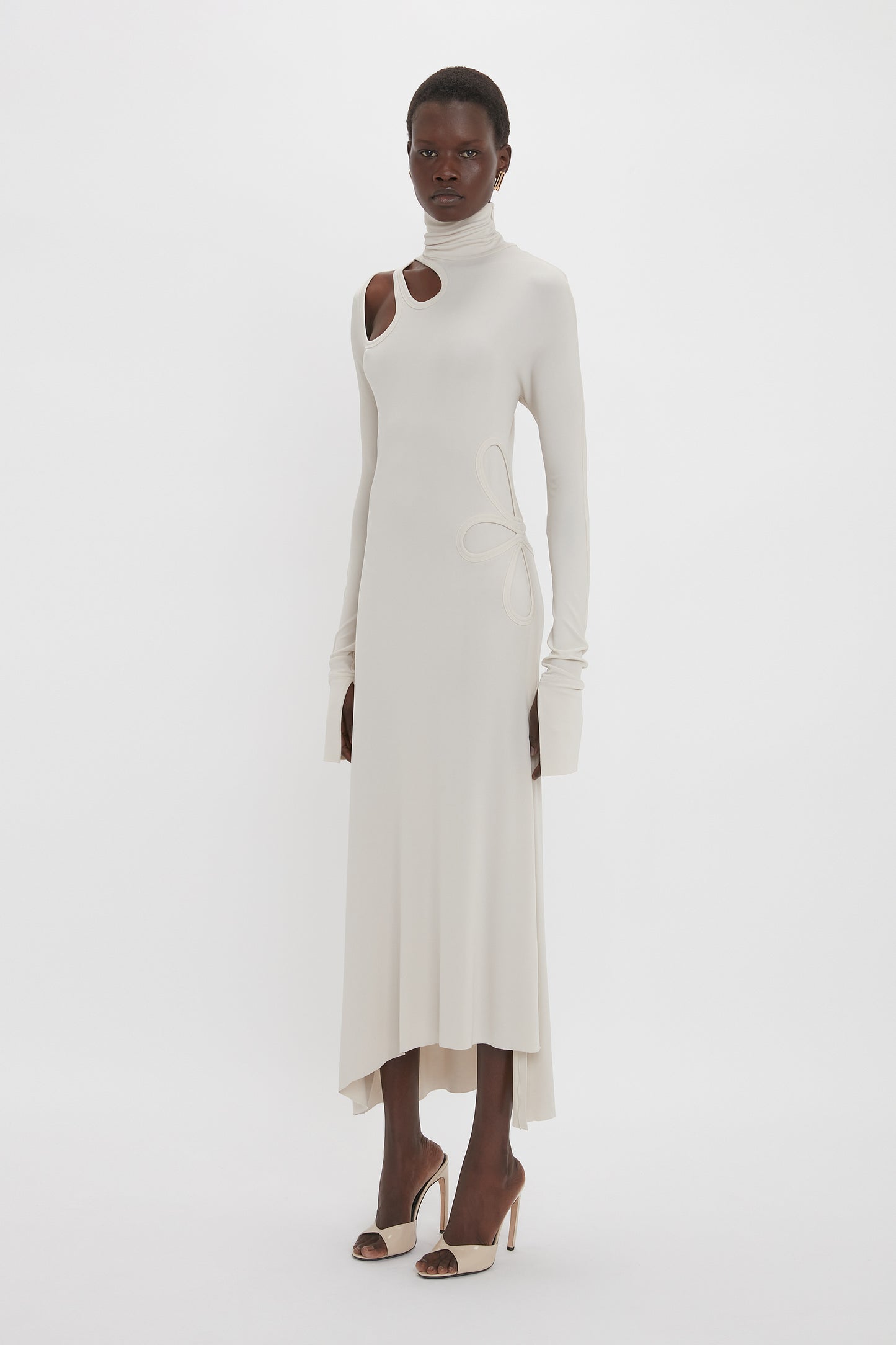 A person stands against a plain white background, wearing a white floor-length Long Sleeve Cut-Out Jersey Midi Dress In Bone by Victoria Beckham, featuring an asymmetric hemline and long sleeves, paired with white high-heeled shoes.