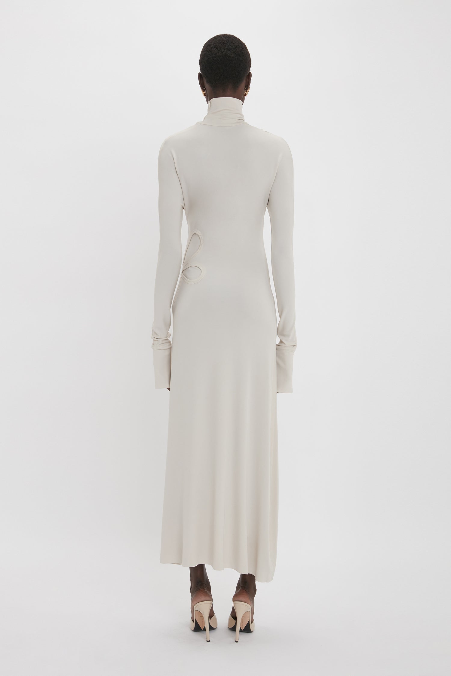 A person with short hair stands facing away, wearing the Long Sleeve Cut-Out Jersey Midi Dress In Bone by Victoria Beckham, featuring a floor-length, cream-colored design and pointed high heels on a plain white background.