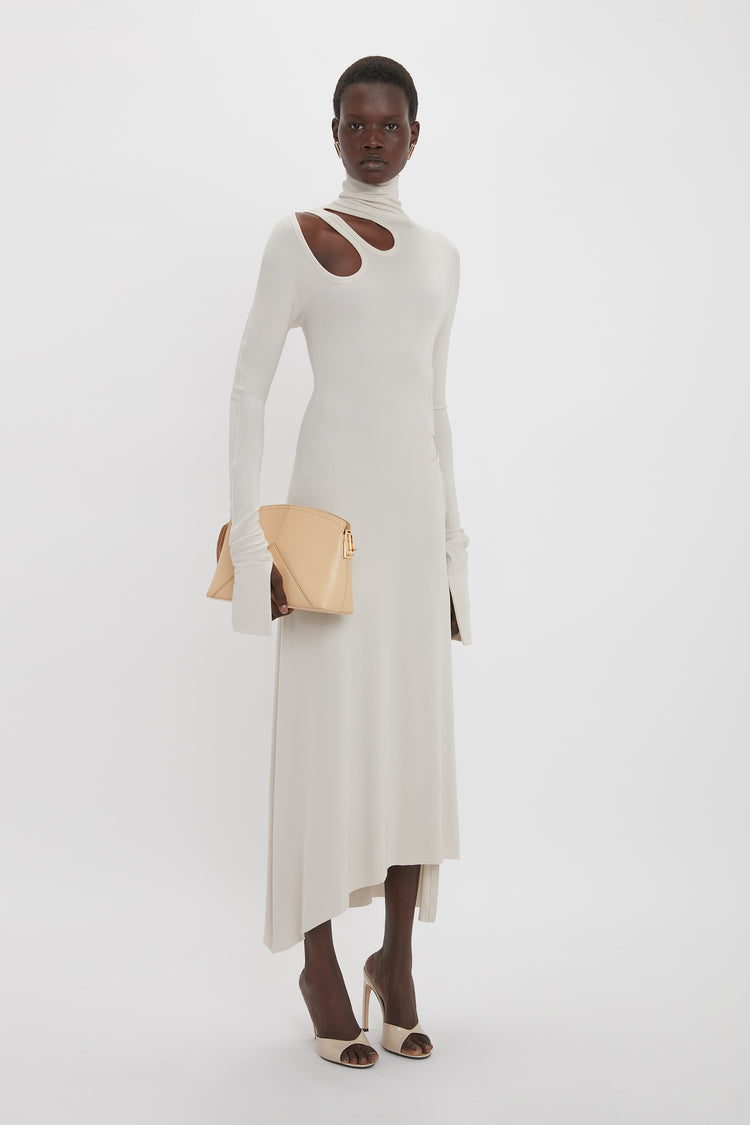 A person stands against a plain background wearing the Victoria Beckham Long Sleeve Cut-Out Jersey Midi Dress In Bone, holding a beige clutch, and matching high-heeled sandals.