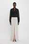 A person stands against a white background wearing the Victoria Beckham Pocket Detail Shirt In Black with tailored design details and a white high-waisted skirt featuring a front slit.