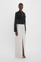 Person standing in a studio setting wearing a lightweight semi-sheer knit, long-sleeve black blouse named the "Pocket Detail Shirt In Black" from Victoria Beckham and a high-waisted, long, white skirt with tailored design details and a front slit.