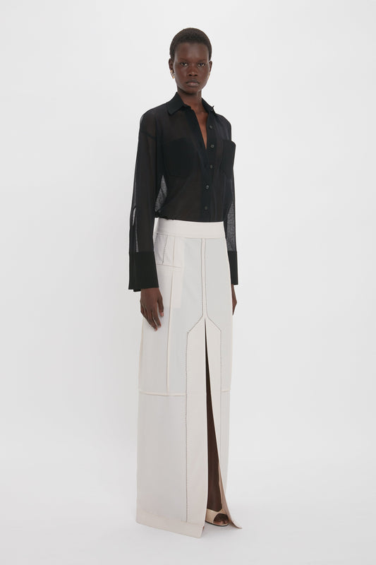 A person stands against a plain background wearing a black, long-sleeve, button-up shirt and an elegant Victoria Beckham Deconstructed Floor-Length Skirt In Bone with a front slit.