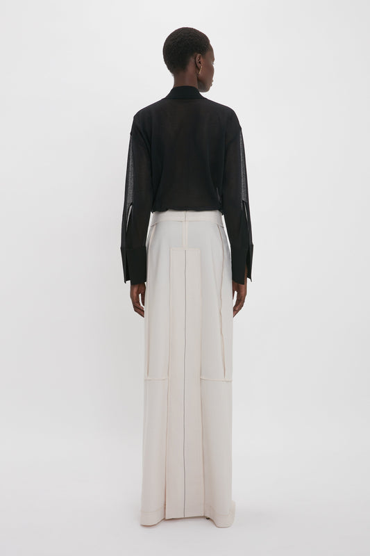 A person stands facing away, showcasing elegant fluidity in a black long-sleeve shirt and the Victoria Beckham Deconstructed Floor-Length Skirt In Bone, against a plain white background.