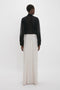 A person stands facing away, showcasing elegant fluidity in a black long-sleeve shirt and the Victoria Beckham Deconstructed Floor-Length Skirt In Bone, against a plain white background.