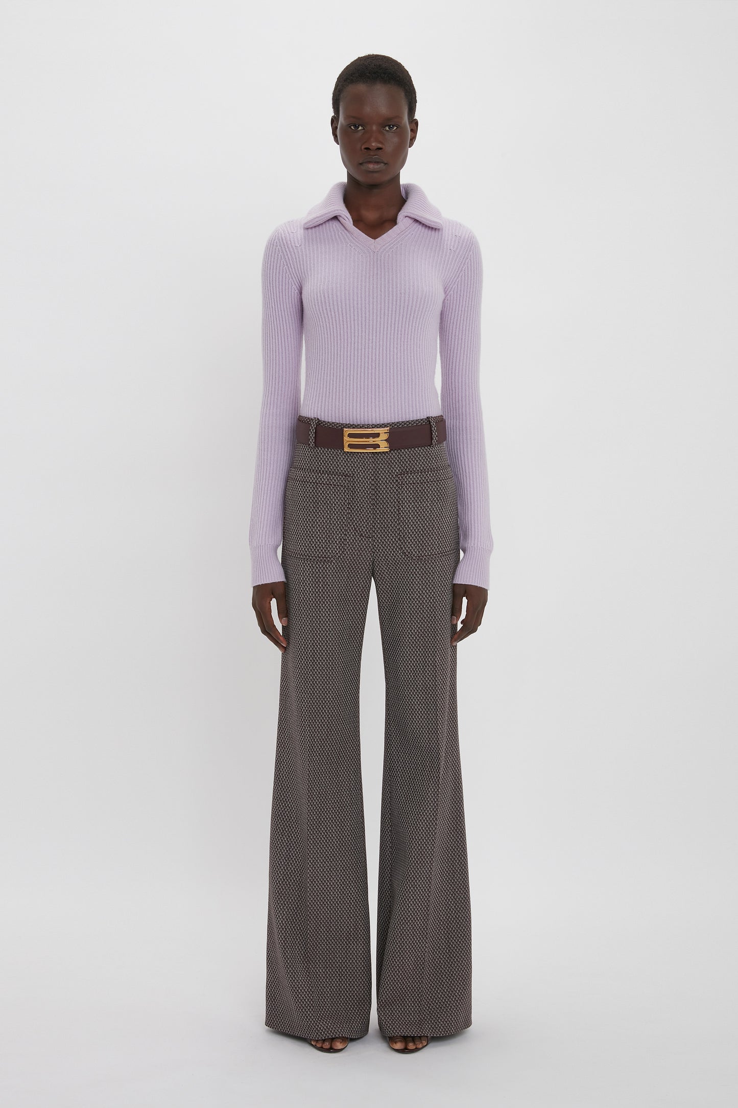 A person stands against a plain white background wearing a light purple Victoria Beckham Double Collared Jumper In Petunia, patterned wide-leg trousers, and a dark belt with a gold buckle.