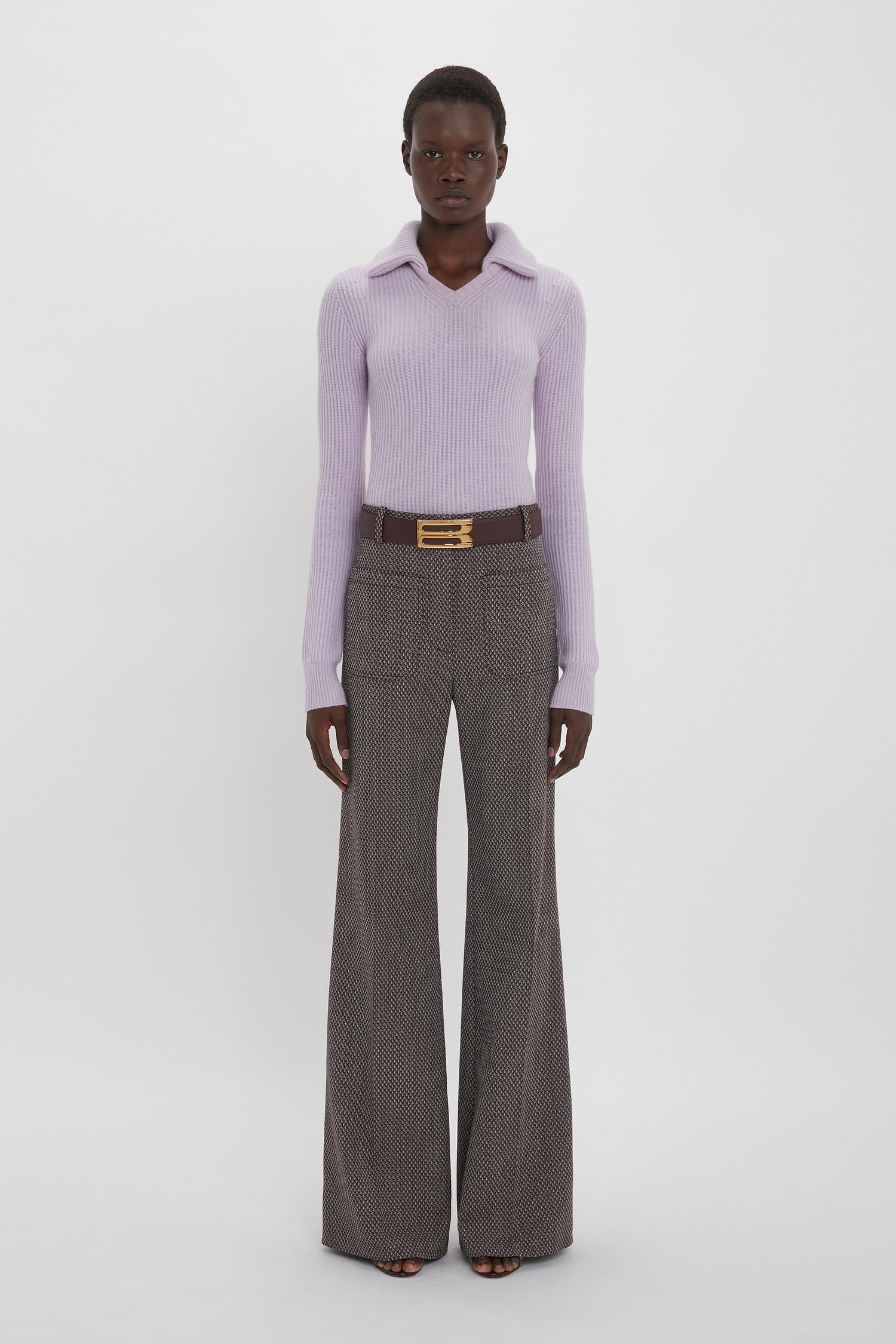 A person stands against a plain white background wearing a light purple Victoria Beckham Double Collared Jumper In Petunia, patterned wide-leg trousers, and a dark belt with a gold buckle.