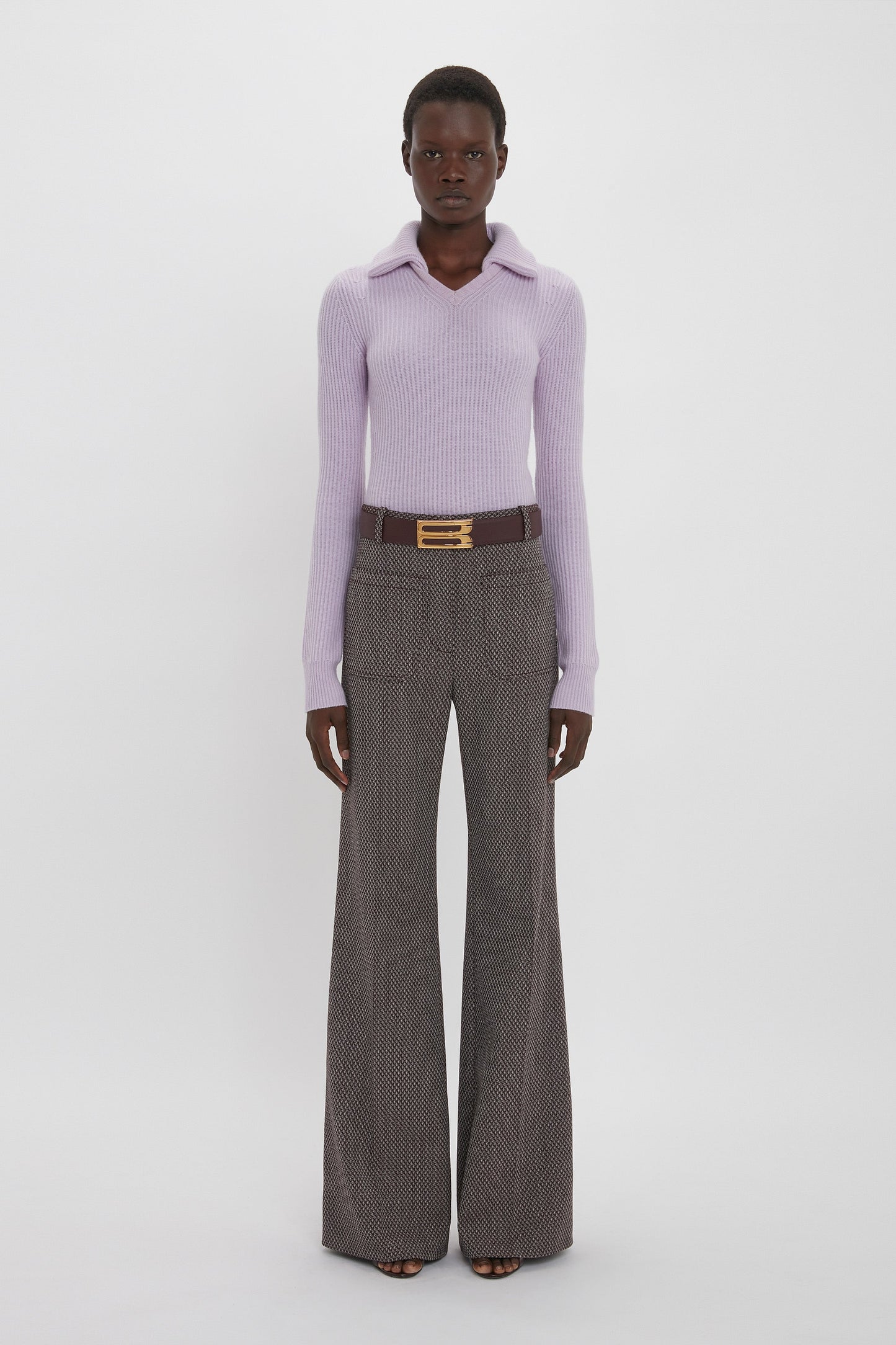 A person stands against a plain background wearing a light purple long-sleeve top and high-waisted Alina Trouser In Tobacco by Victoria Beckham, complemented by a brown belt with a large gold buckle.