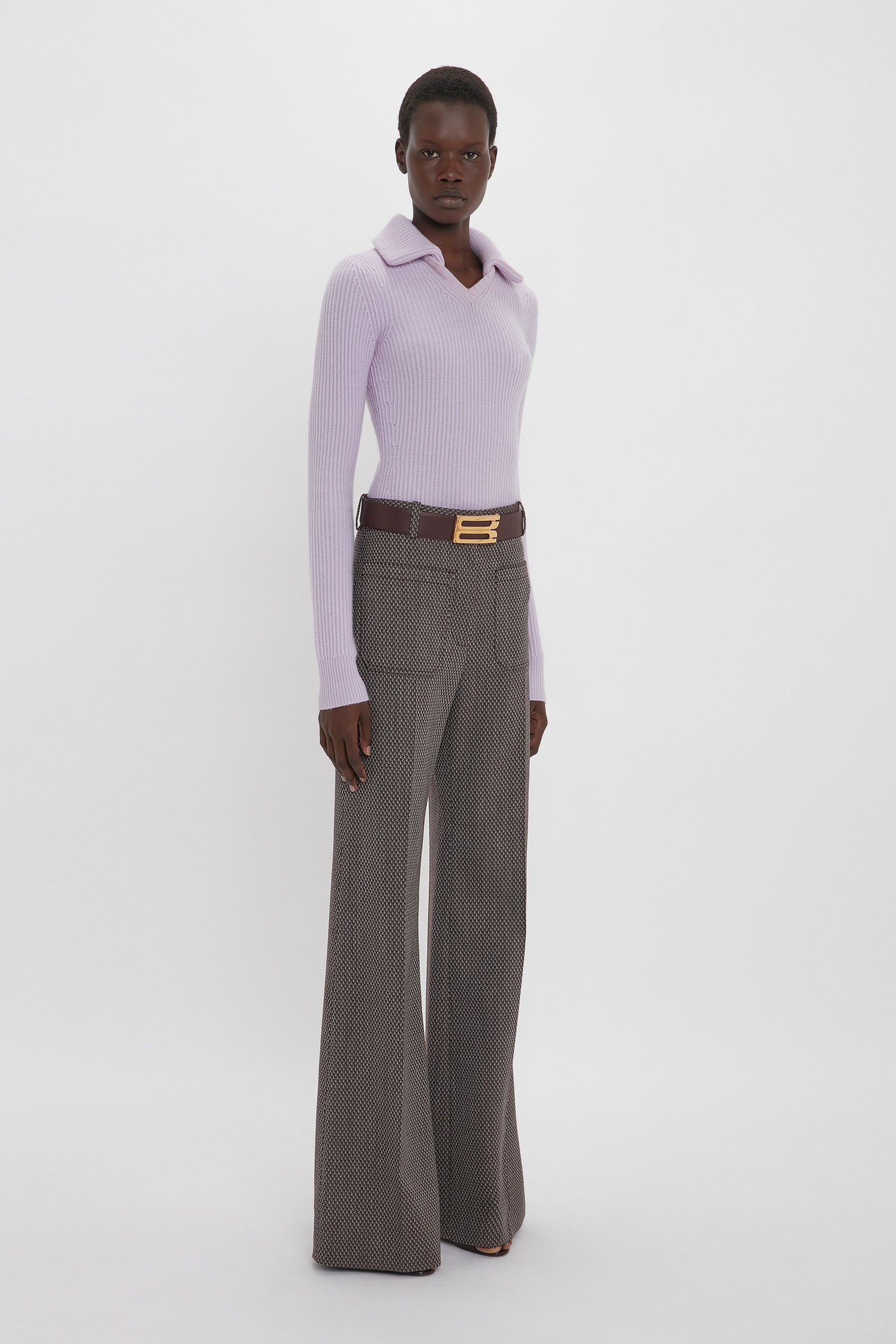 A person stands against a plain background, wearing a Victoria Beckham Double Collared Jumper In Petunia and high-waisted, wide-leg brown pants with a belt.