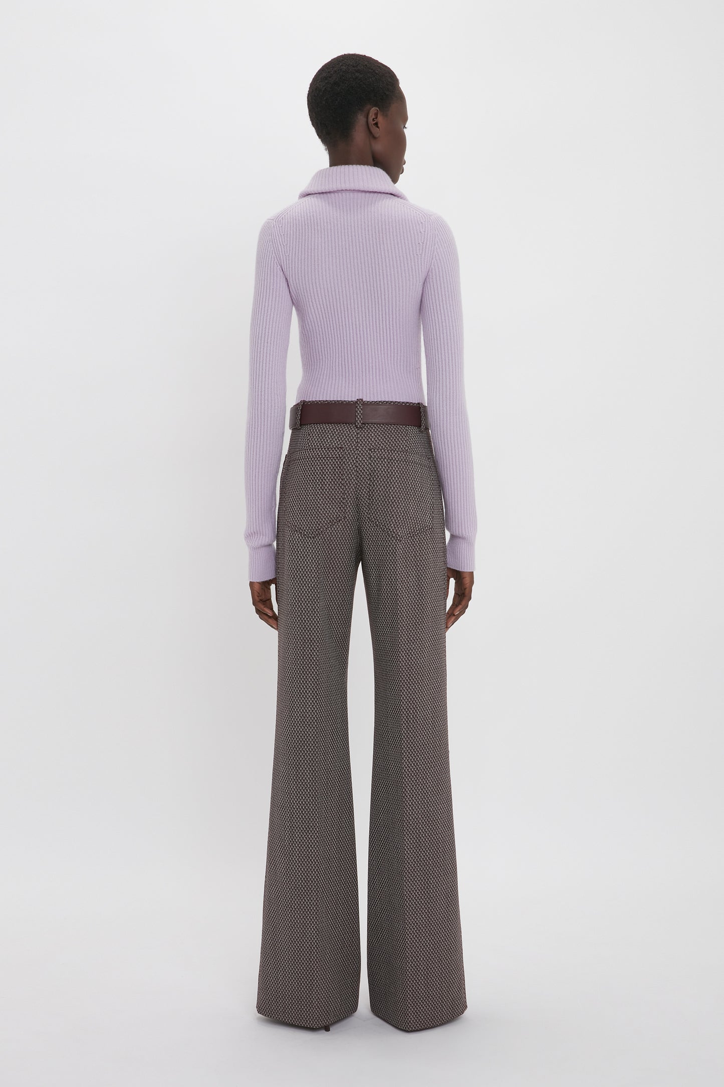 A person stands with their back facing the camera, wearing a Victoria Beckham Double Collared Jumper In Petunia and high-waisted brown wide-legged pants.