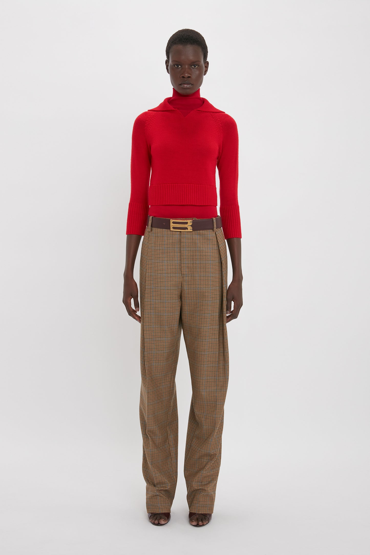 A person stands against a plain background, wearing a Double Layer Top In Deep Red by Victoria Beckham and high-waisted brown plaid trousers with a belt.