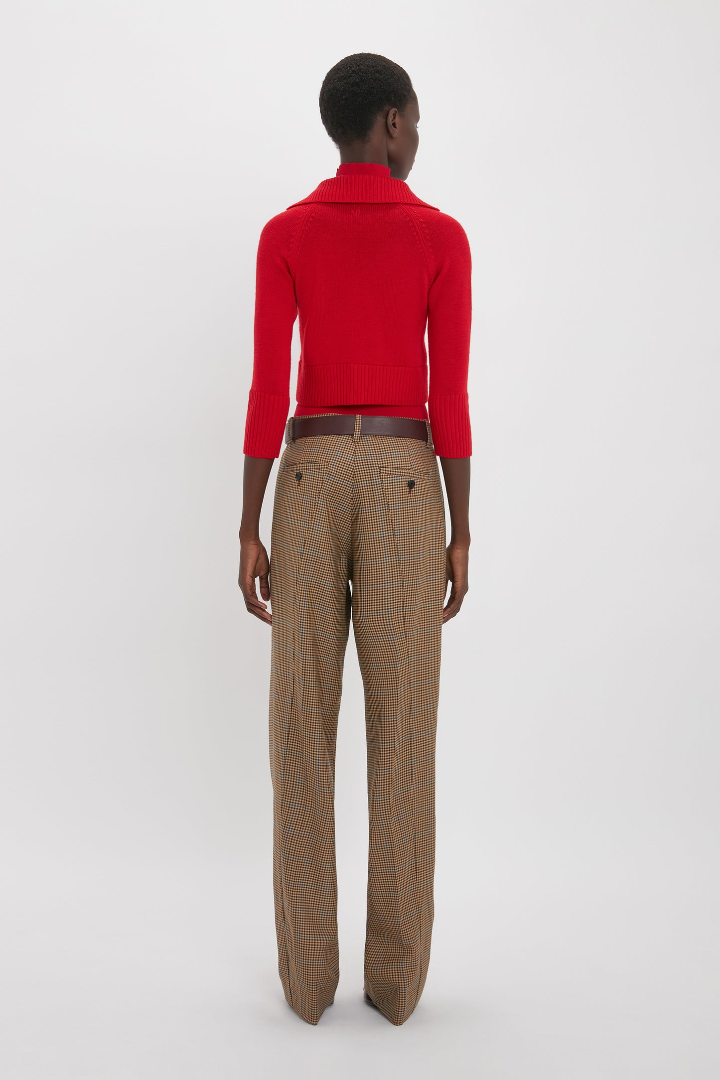Person standing and facing away from the camera, wearing a Victoria Beckham Double Layer Top In Deep Red and high-waisted, wide-leg plaid pants.