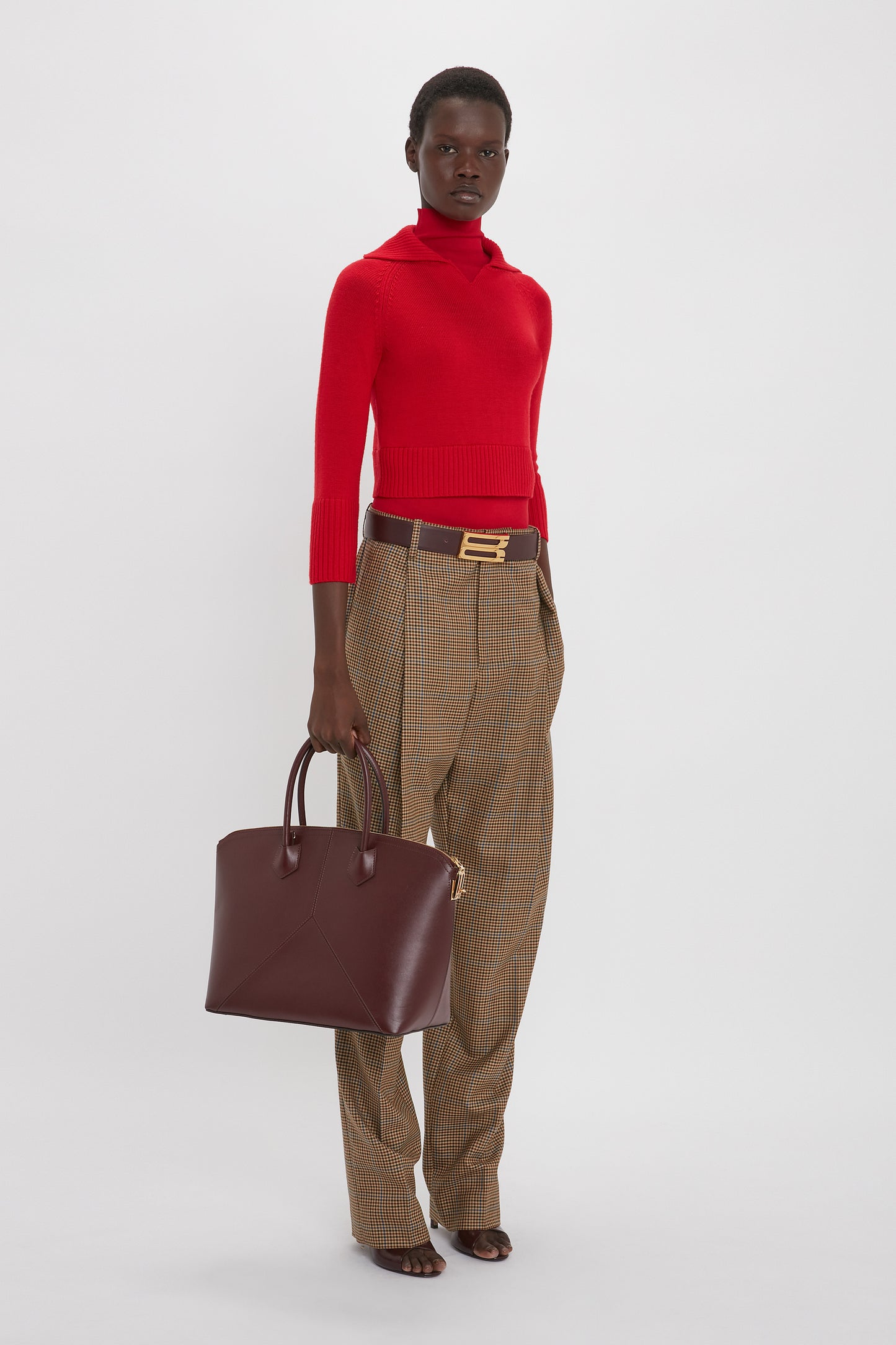 Person wearing a Victoria Beckham Double Layer Top In Deep Red, brown high-waisted pants with a gold belt, and holding a dark brown handbag stands against a plain white background.