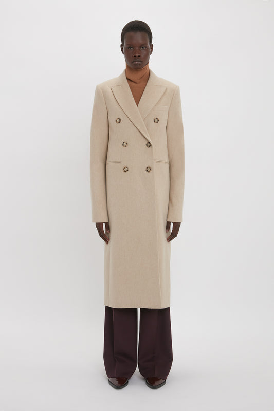 A black model wearing a Victoria Beckham Tailored Slim Coat In Bone over a brown sweater, paired with dark brown trousers and shoes, standing against a white background.