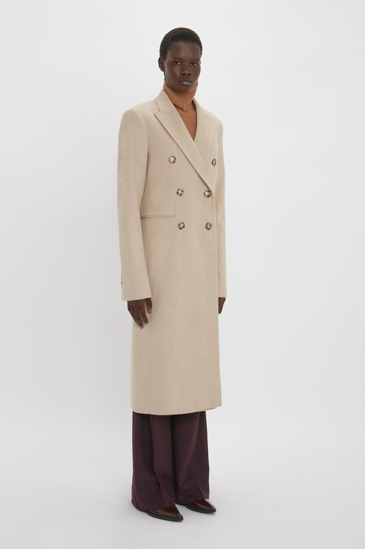 A black man wearing a long beige double-breasted overcoat by Victoria Beckham and dark trousers, standing against a plain white background.