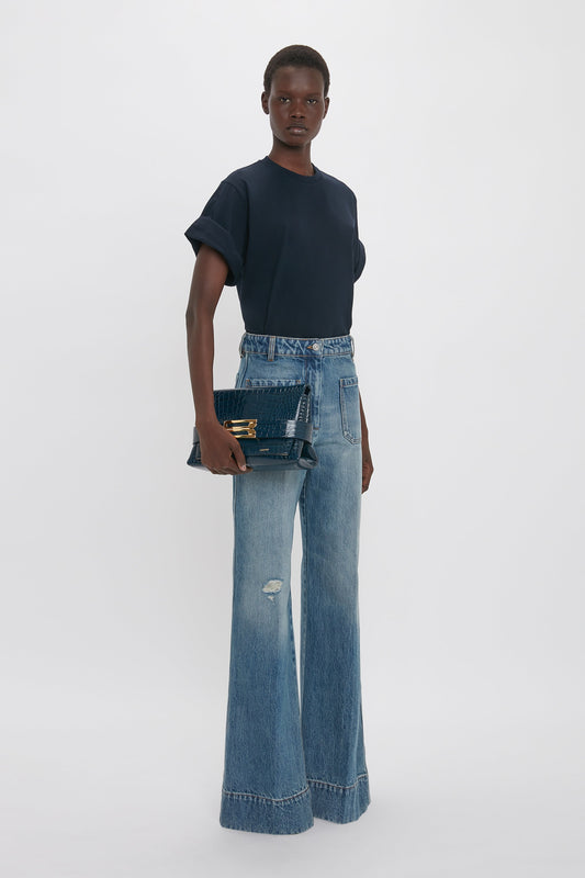 A person stands against a plain backdrop wearing a navy blue t-shirt, high-waisted flared jeans, and holding a Victoria Beckham B Pouch Bag In Croc Effect Midnight Blue Leather.