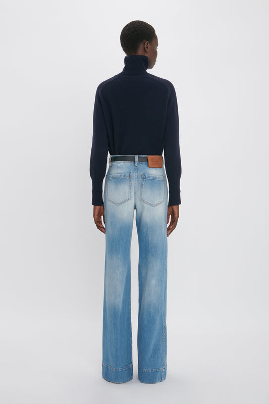 A person stands facing away, wearing a dark turtleneck sweater and light blue wide-leg vintage denim jeans with a visible brown leather patch on the back waistband. The background is plain white. They are wearing the Alina Jean In Light Summer Wash by Victoria Beckham.