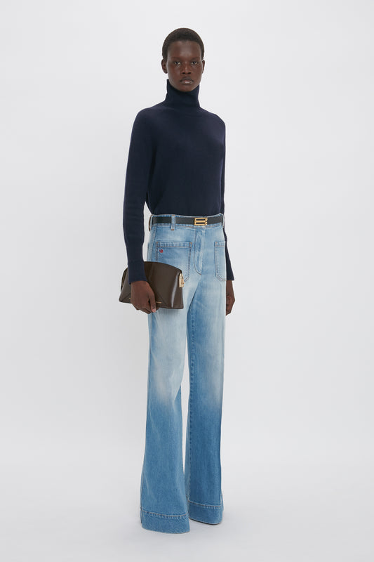 A person stands against a plain background, wearing a navy turtleneck sweater, Victoria Beckham Alina Jean In Light Summer Wash, and holding a brown clutch.