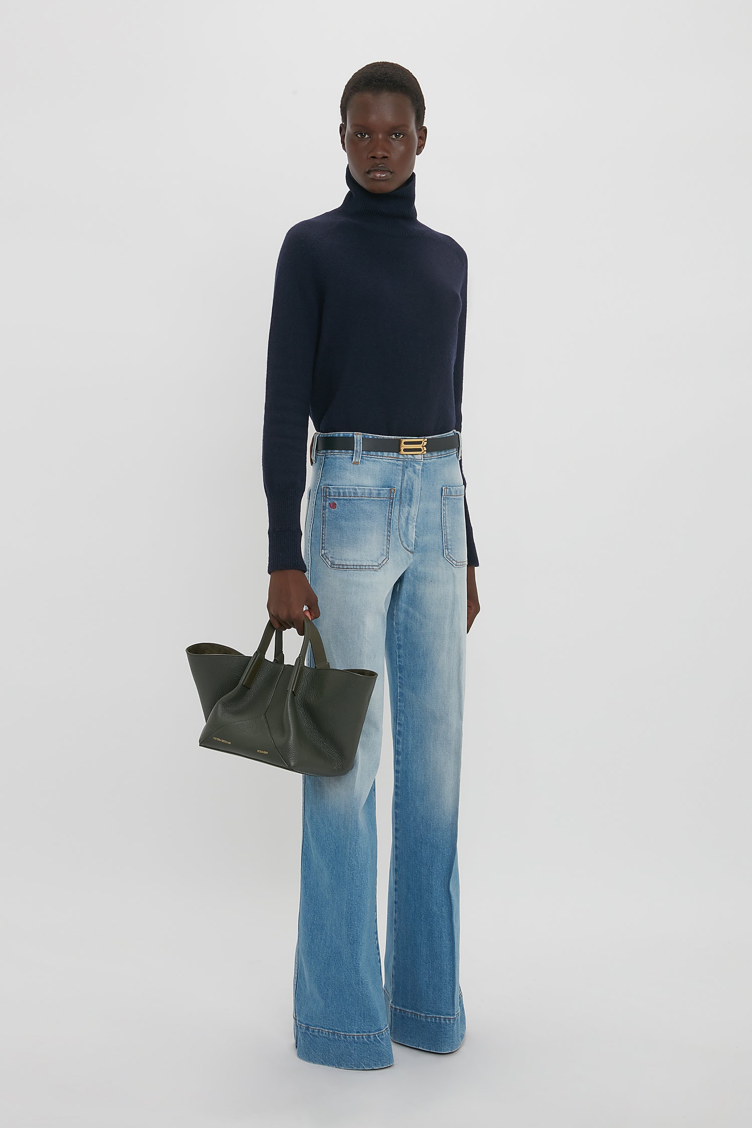 Person wearing a navy turtleneck, light blue wide-leg jeans, and holding a luxurious Victoria Beckham W11 Mini Tote Bag In Loden Leather stands against a plain white background.