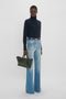 Person wearing a navy turtleneck, light blue wide-leg jeans, and holding a luxurious Victoria Beckham W11 Mini Tote Bag In Loden Leather stands against a plain white background.