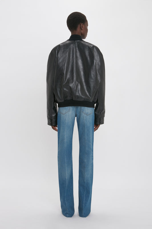 A person stands facing away, wearing a Victoria Beckham Leather Varsity Jacket In Black and blue jeans. The background is plain white.