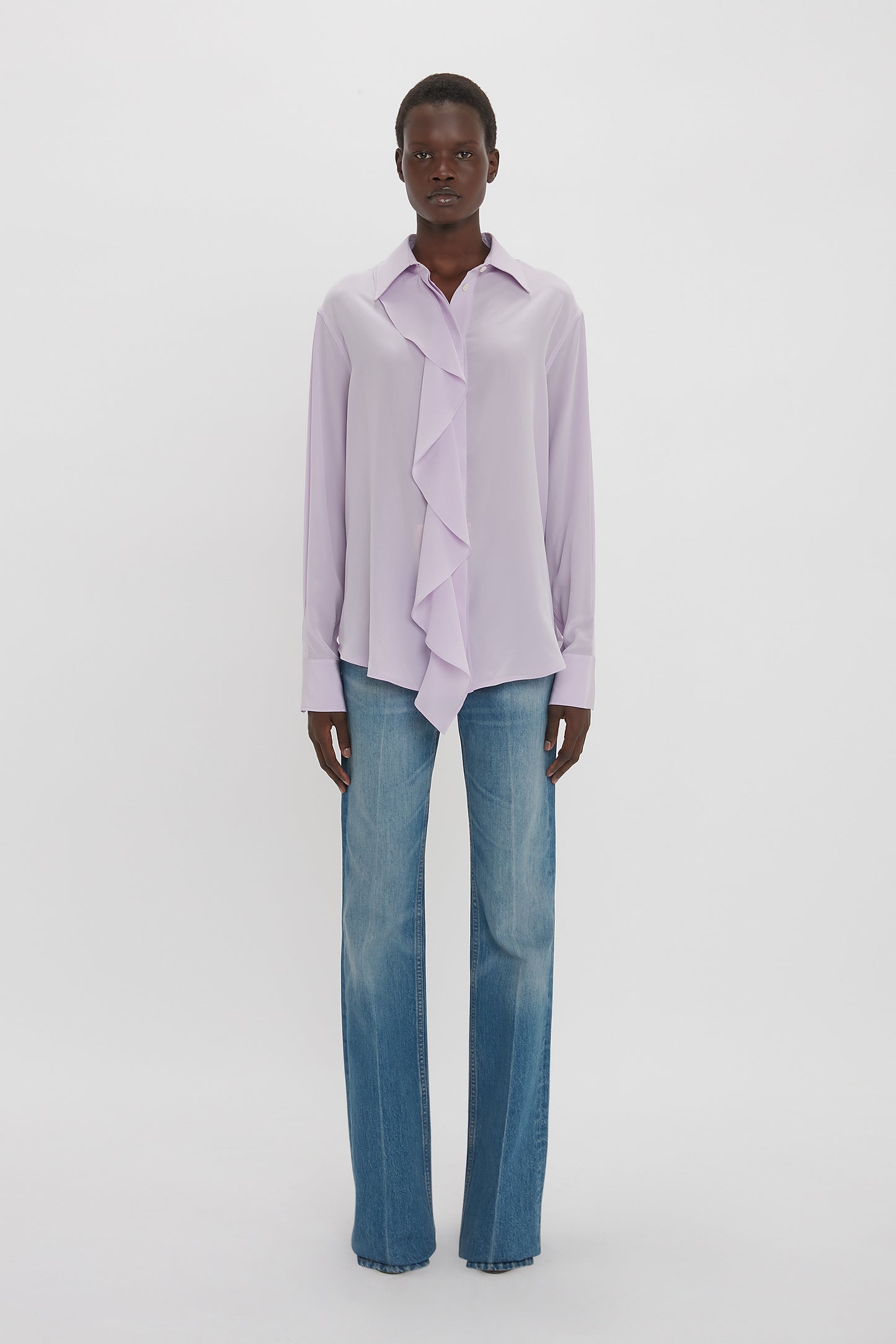 Person wearing an ethereal shade of Victoria Beckham Asymmetric Ruffle Blouse In Petunia paired with wide-leg blue jeans, standing against a plain white background.