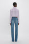 Rear view of a person wearing a Victoria Beckham asymmetric ruffle blouse in petunia and blue jeans, standing against a white background.