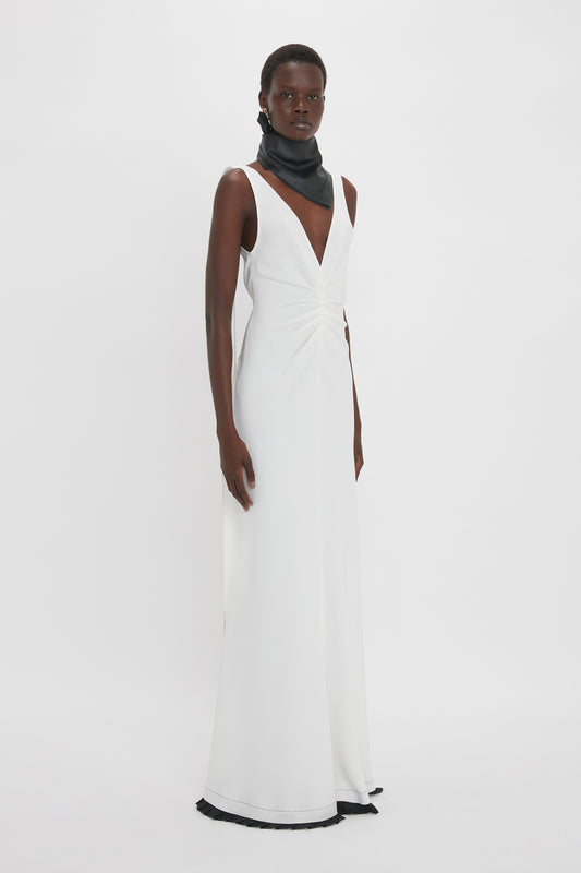 A woman stands on a white background wearing the Victoria Beckham Exclusive V-Neck Gathered Waist Floor-Length Gown in Ivory with a sleeveless design, deep V-neck, and black scarf around her neck, perfectly highlighting her hourglass silhouette.