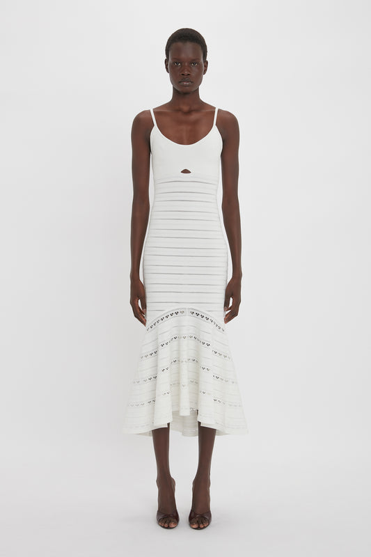 A person stands facing forward wearing a white Cut-Out Detail Cami Dress In White by Victoria Beckham with thin shoulder straps and a flared silhouette. The background is plain and white.