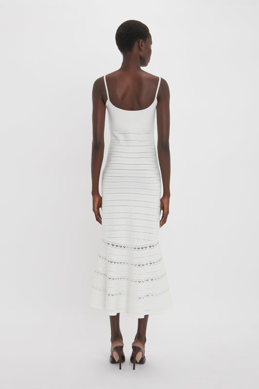 A person stands facing away from the camera, wearing a sleeveless, ankle-length white Cut-Out Detail Cami Dress In White by Victoria Beckham with delicate eyelet detailing near the hem and strappy high-heeled sandals.