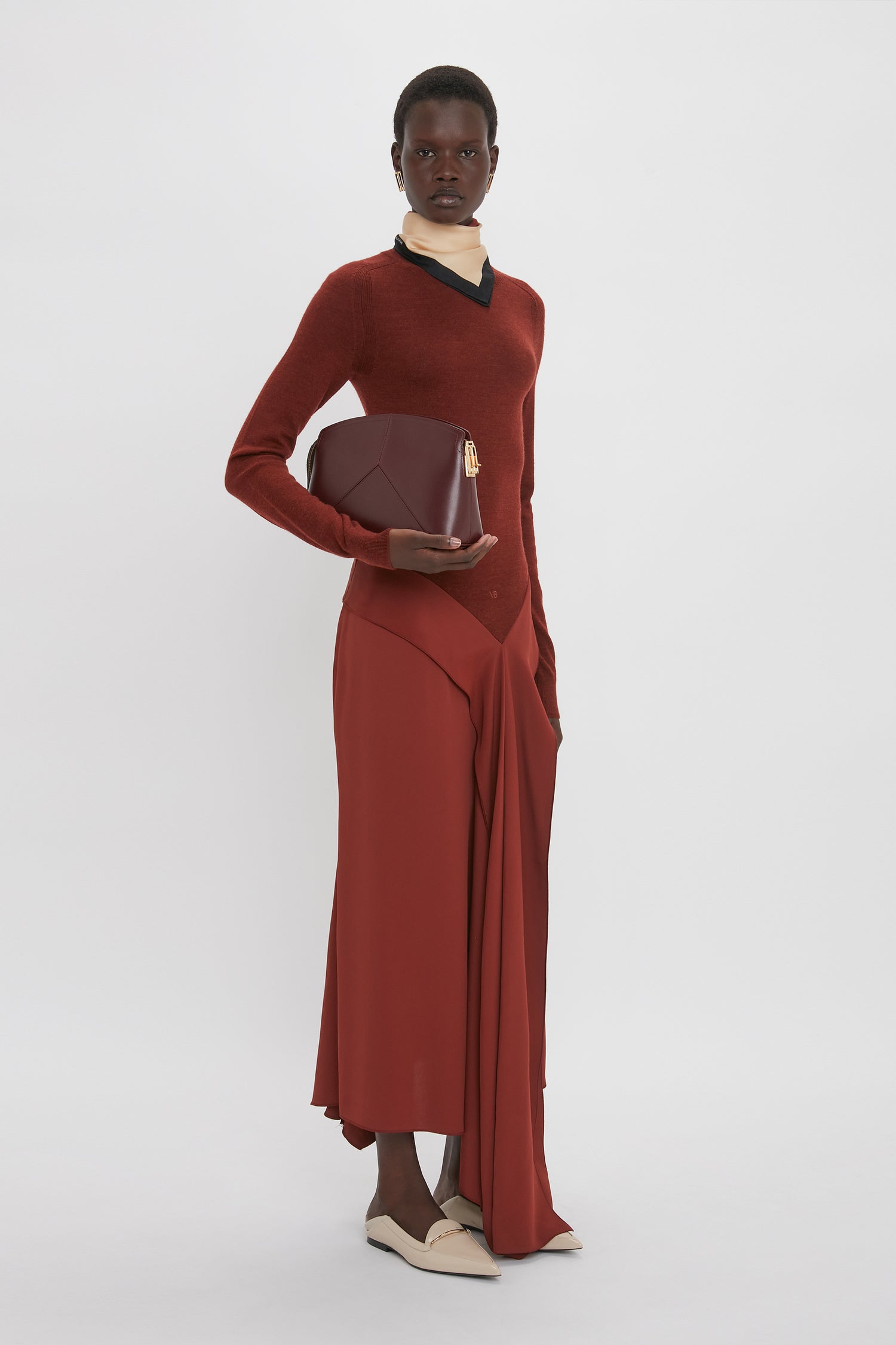 Person wearing a red long-sleeve High Neck Tie Detail Dress In Russet by Victoria Beckham, holding a dark red clutch, standing against a plain white background.