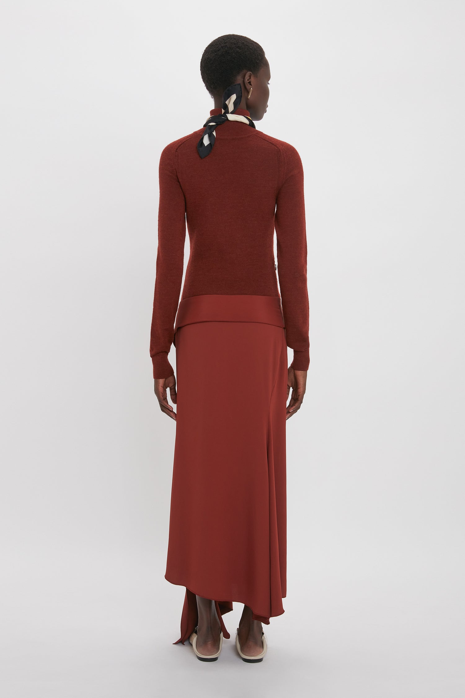 A person wearing a long-sleeve red top and a matching long skirt, reminiscent of Victoria Beckham's High Neck Tie Detail Dress In Russet collection, stands facing away from the camera against a plain background.