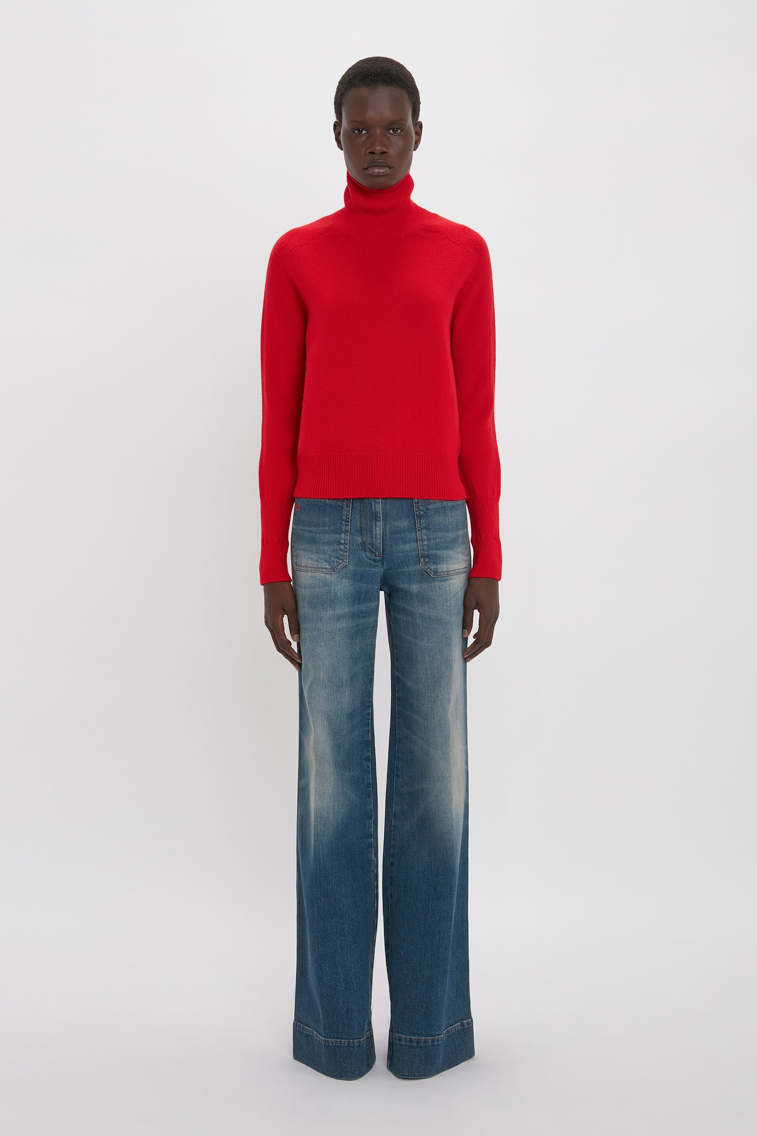 A man wearing a Victoria Beckham red lambswool polo neck jumper and blue jeans standing against a plain white background.