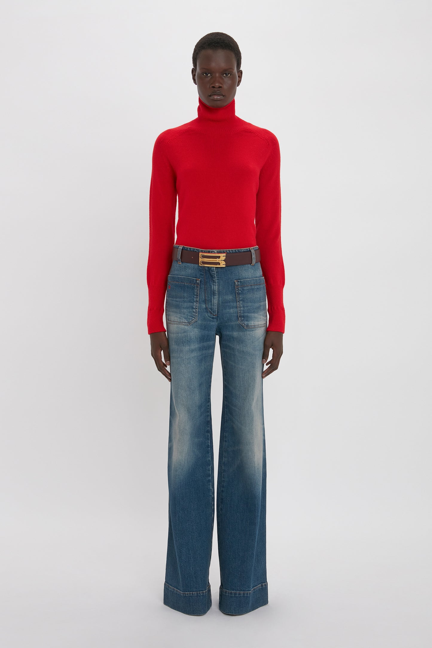 A young black woman in a fitted Victoria Beckham lambswool polo neck jumper in red and blue flared jeans, accessorized with a brown belt. She stands against a plain white background.