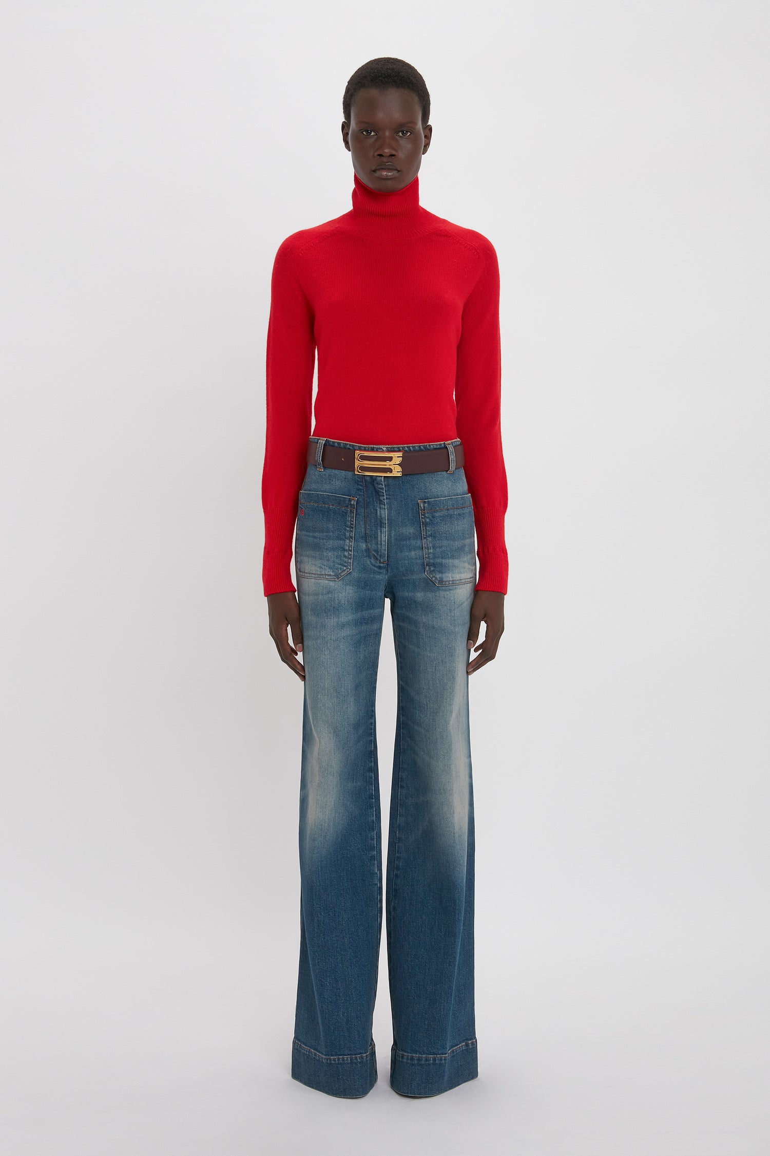 A young black woman in a fitted Victoria Beckham lambswool polo neck jumper in red and blue flared jeans, accessorized with a brown belt. She stands against a plain white background.