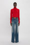 A young black woman in a fitted Victoria Beckham lambswool polo neck jumper in red and blue flared jeans, accessorized with a brown belt. She stands against a plain white background.