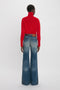 Rear view of a woman wearing a Victoria Beckham red lambswool polo neck jumper and blue wide-leg jeans, standing against a white background.