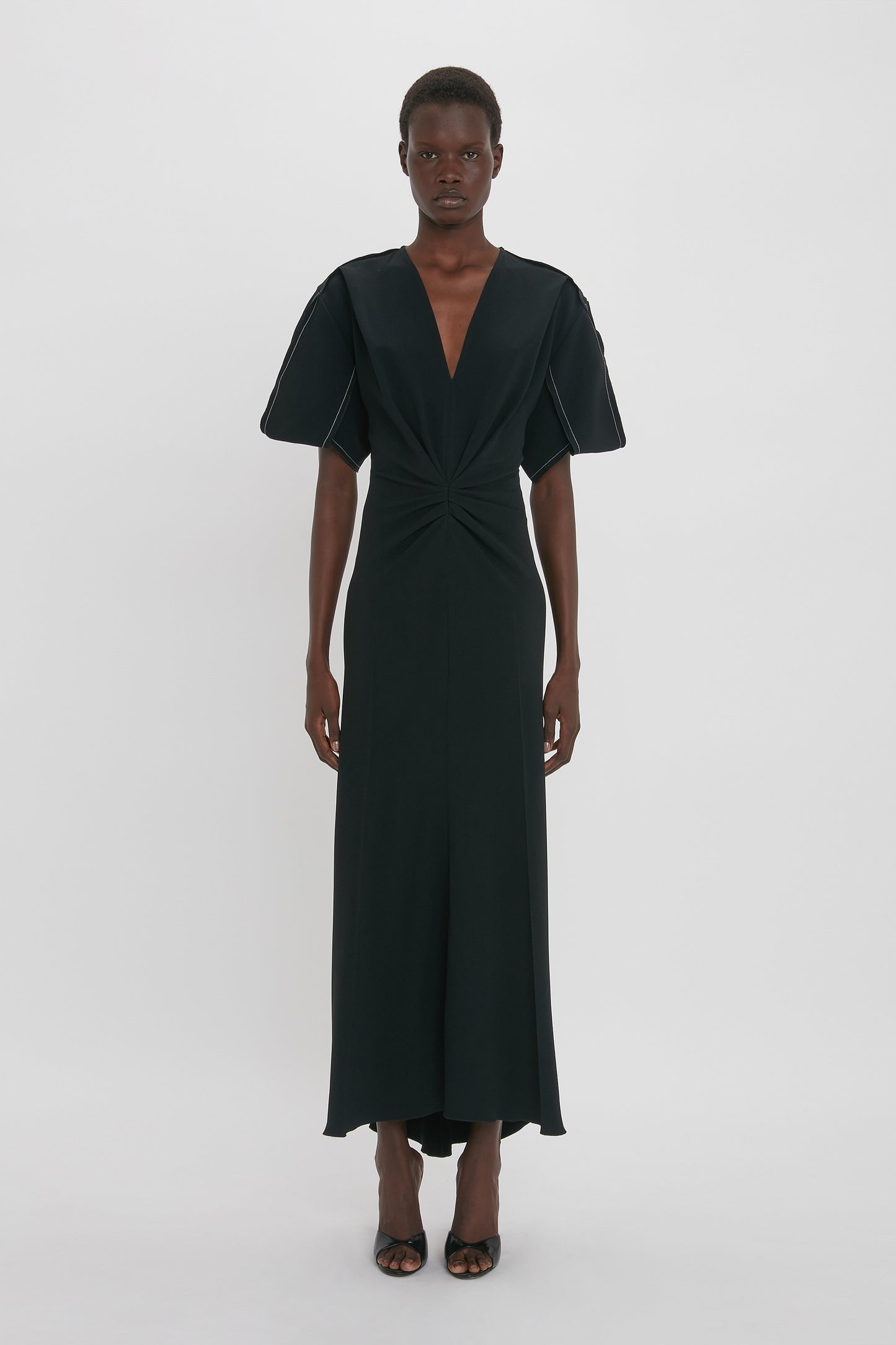 A person stands in a long, black Gathered V-Neck Midi Dress in Black by Victoria Beckham with short sleeves and a deep V-neck, made from figure-flattering stretch fabric, against a plain white background. They wear black high-heeled sandals, adding a contemporary edge to the look.